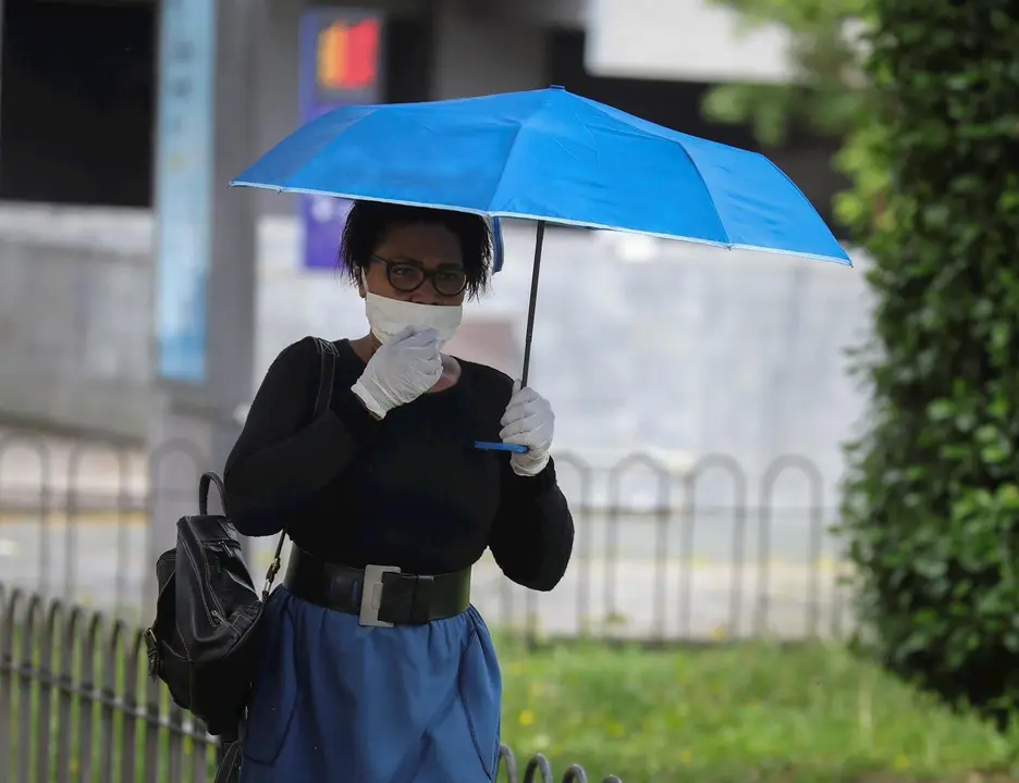 Una mujer protegida con mascarilla y guantes se resguarda de la lluvia bajo su parag&uuml;as en la quinta semana del estado de alarma por el coronavirus, Covid-19. En Sevilla (Andaluc&iacute;a, Espa&ntilde;a), a 15 de abril 2020.
