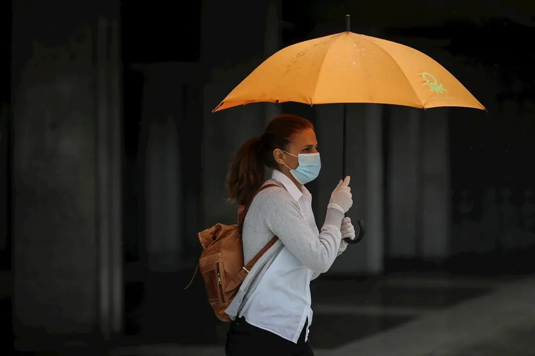 Una mujer protegida con mascarilla y guantes se resguarda de la lluvia bajo su paraguas en la quinta semana del estado de alarma por el coronavirus, Covid-19. En Sevilla (Andaluc&iacute;a, Espa&ntilde;a), a 15 de abril 2020.