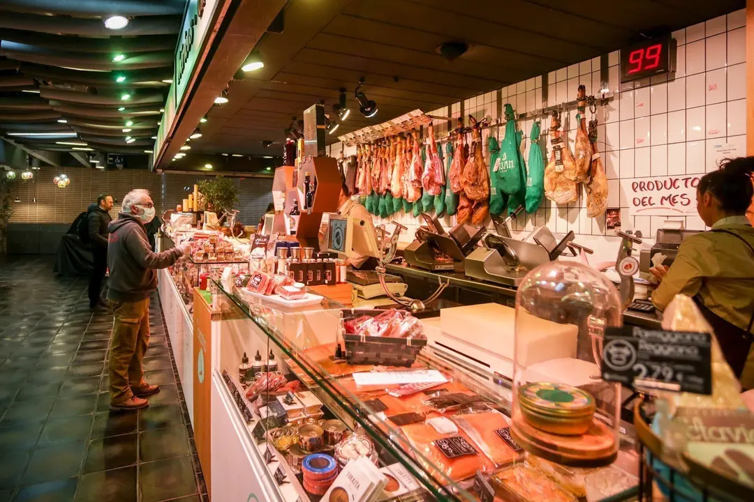 Varios clientes hacen la compra en un puesto de carne y embutidos en el mercado de San Ant&oacute;n, que permanece abierto y anuncia que tambi&eacute;n atiende a domicilio, en Madrid (Espa&ntilde;a), a 18 de marzo de 2020.