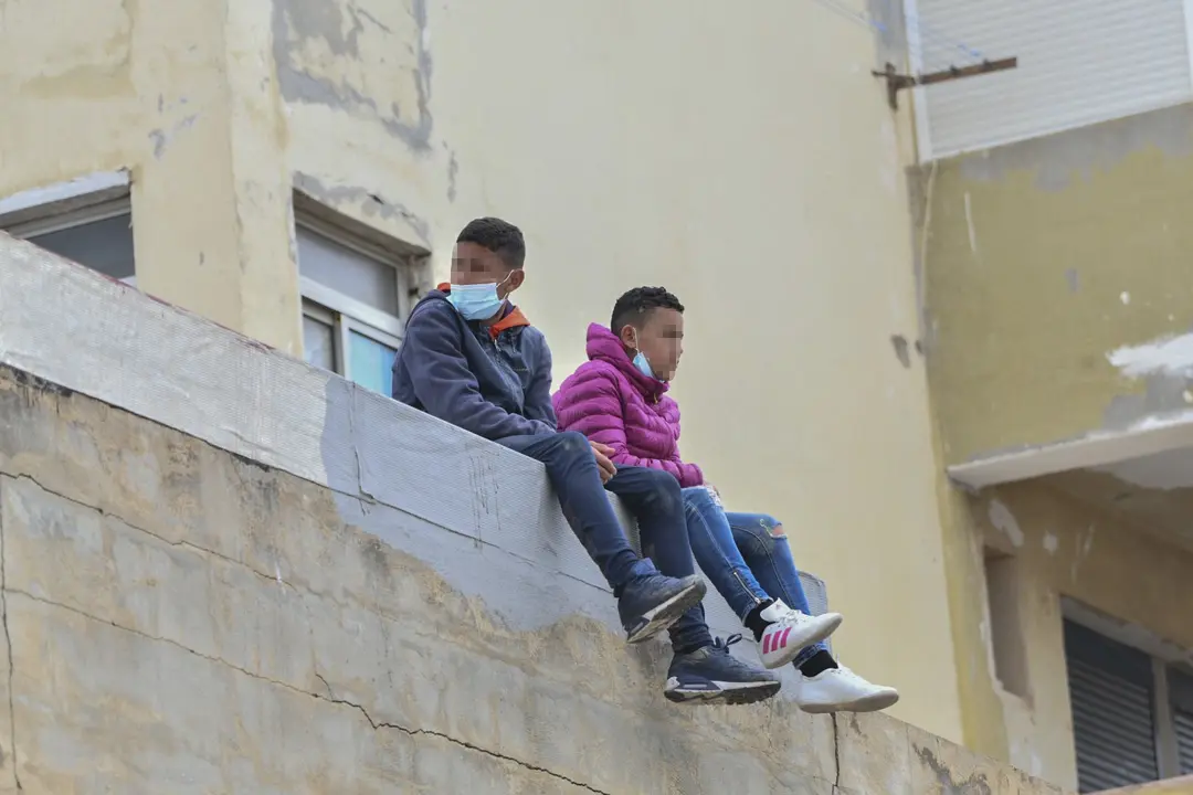 Dos ni&ntilde;os sentados sobre un muro en la nave de primera acogida del pol&iacute;gono del Tarajal en Ceuta (Espa&ntilde;a)