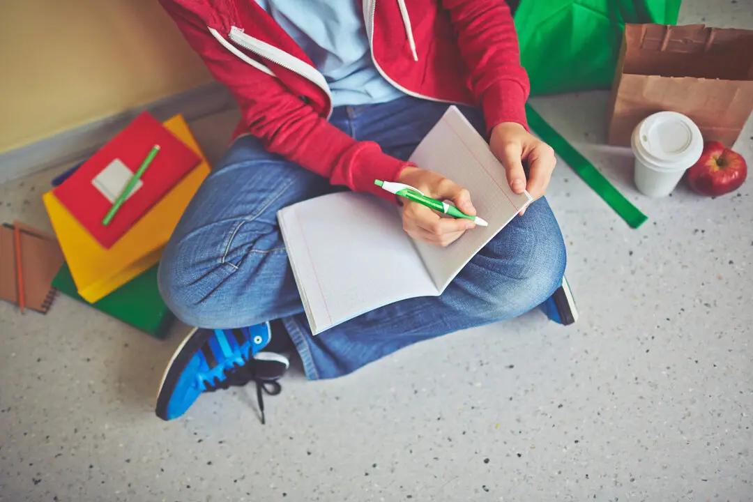Schoolboy in casualwear making notes in exercise-book while sitting on the floor