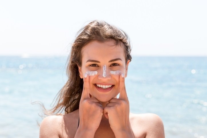 Beautiful young woman is posing at the camera with suncream on her face over the sea background.