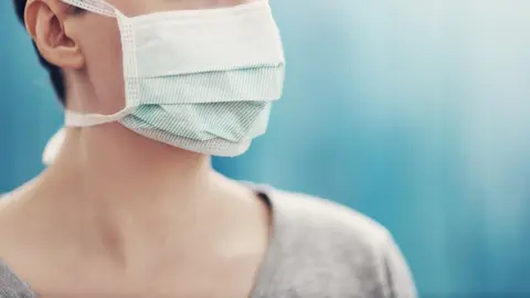 young woman in medical face protection mask indoors on blue background. Sick person