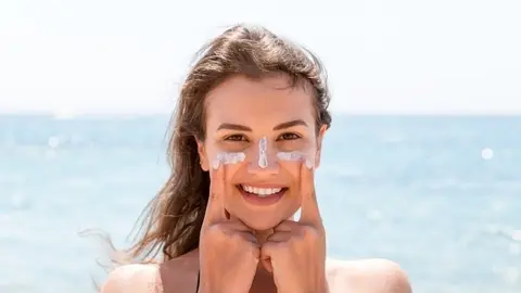 Beautiful young woman is posing at the camera with suncream on her face over the sea background.