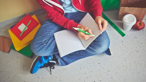 Schoolboy in casualwear making notes in exercise-book while sitting on the floor